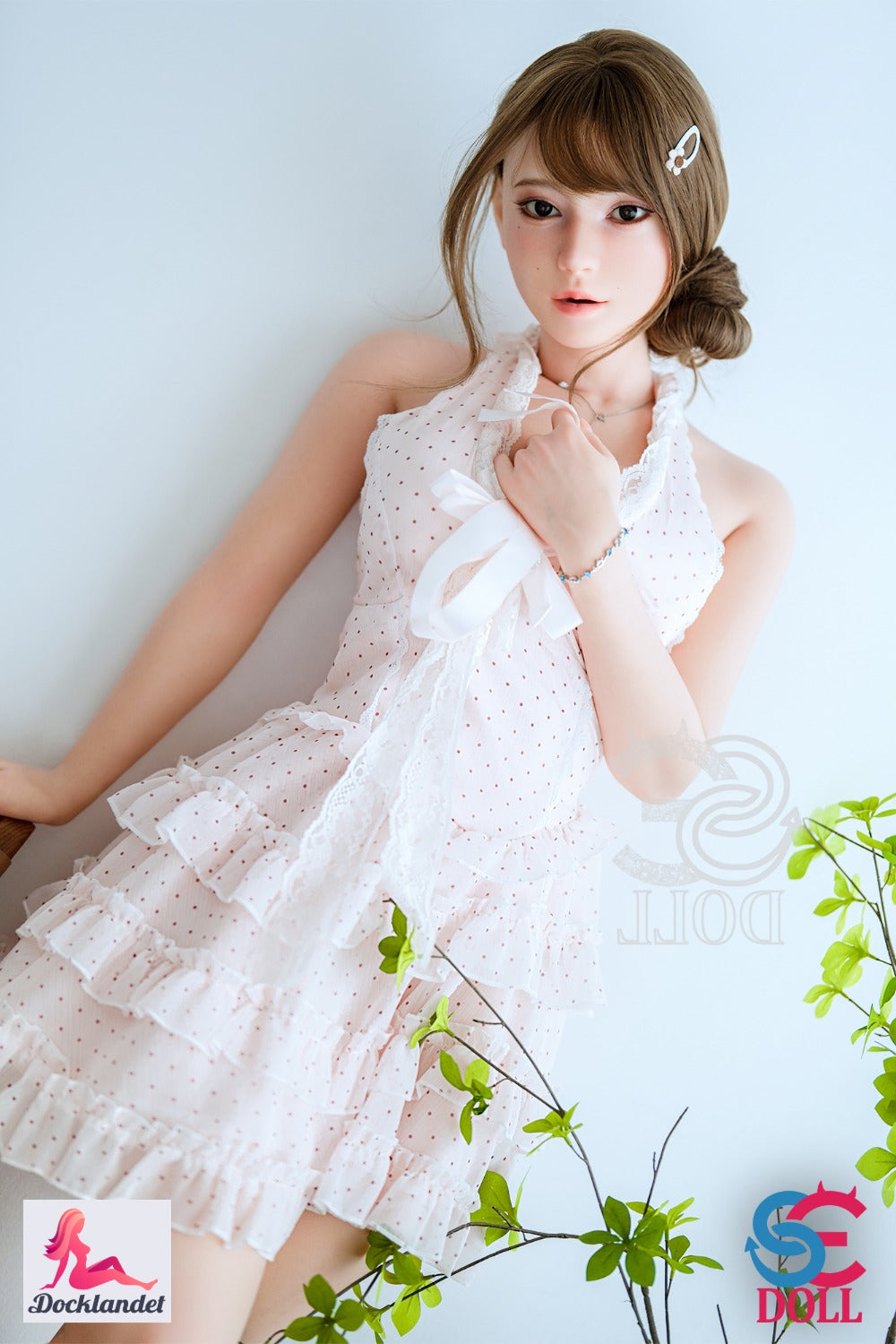Maya.B no SE-Doll 163 cm ar C-kausu, brūnu frizūru un baltiem mežģīņu svārkiem, reālistiska pilna silikona lelle sēž uz krēsla.