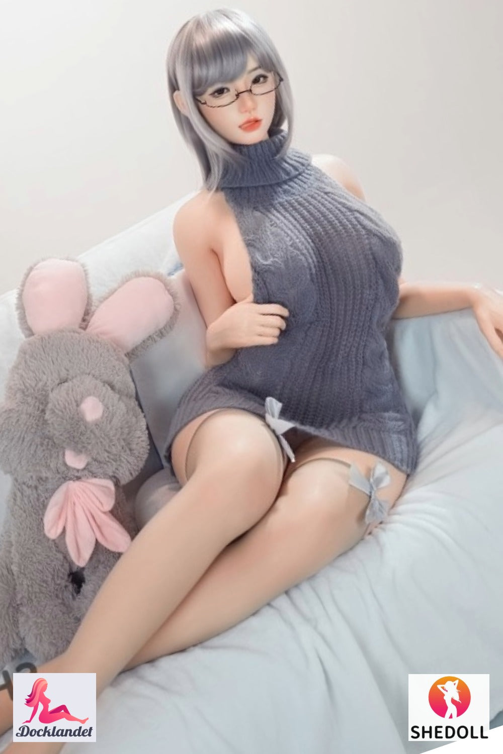 Shedoll seksa lelle ar īsu pelēku matu sakārtojumu un brillēm pelēkā trikotāžas kleitā sēž uz dīvāna blakus trusis izbāztajam dzīvniekam - reālistiska silikona lelle no plkst. Docklandet